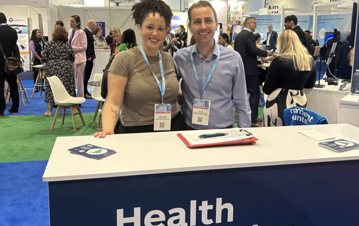 A male and female member of Health Innovation Network staff smile from behind a reception desk at an event, ready to help delegates.