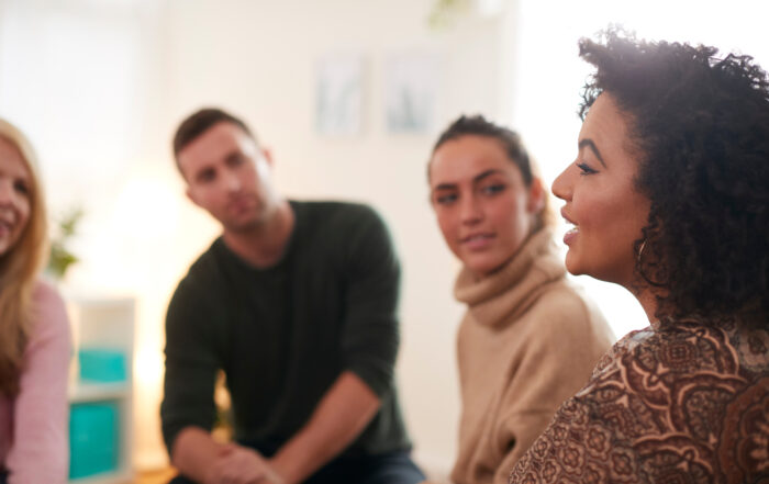 An older lady is talking openly to a group of younger people, smiling as she talks passionately at the support group. The group are looking eagerly at her, in a community space.